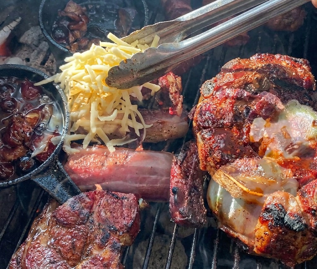 Tongs dropping shredded yellow and white cheese onto a grilled meat cup filled with a potato and blueberry sauce.