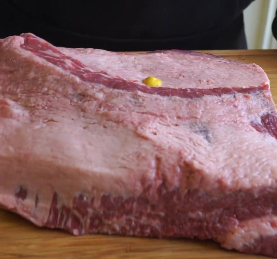 A stream of yellow mustard hitting the surface of a large piece of raw beef brisket on a wooden board.