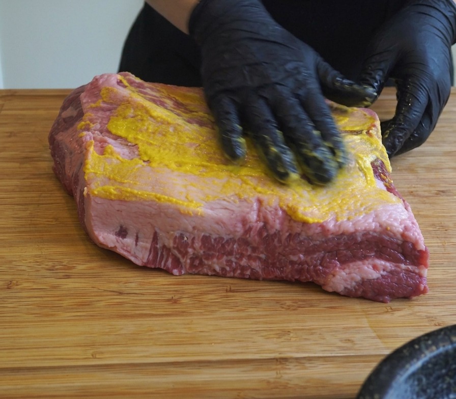Hands in black gloves spreading yellow mustard and salt onto a large brisket on a wooden cutting board.