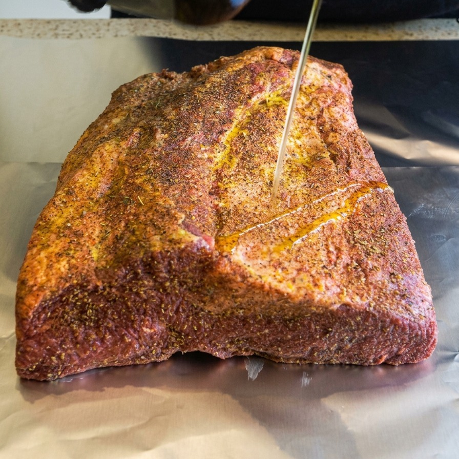 A thin stream of olive oil being drizzled onto a spice-coated beef brisket resting on silver foil.