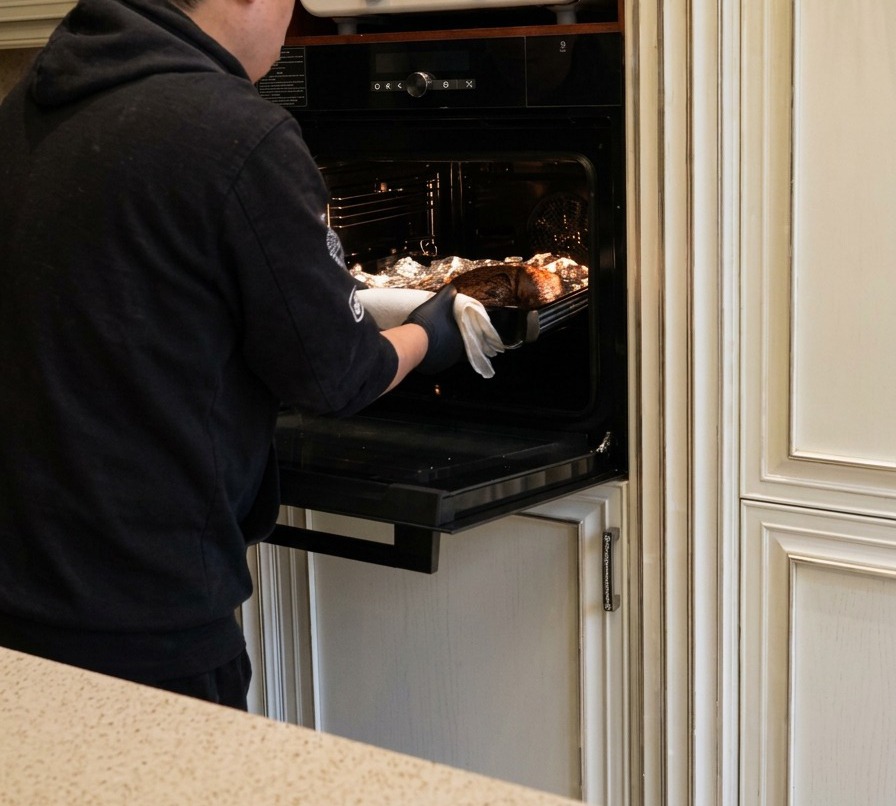 A person in a black hoodie removing a baking tray from an oven, displaying a large, dark-brown roasted beef brisket.