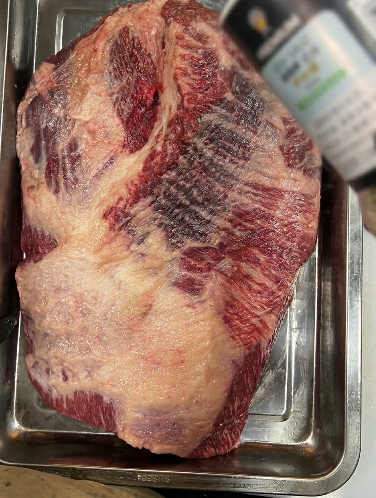 Coarse sea salt being sprinkled onto a large piece of raw brisket sitting in a stainless steel tray.