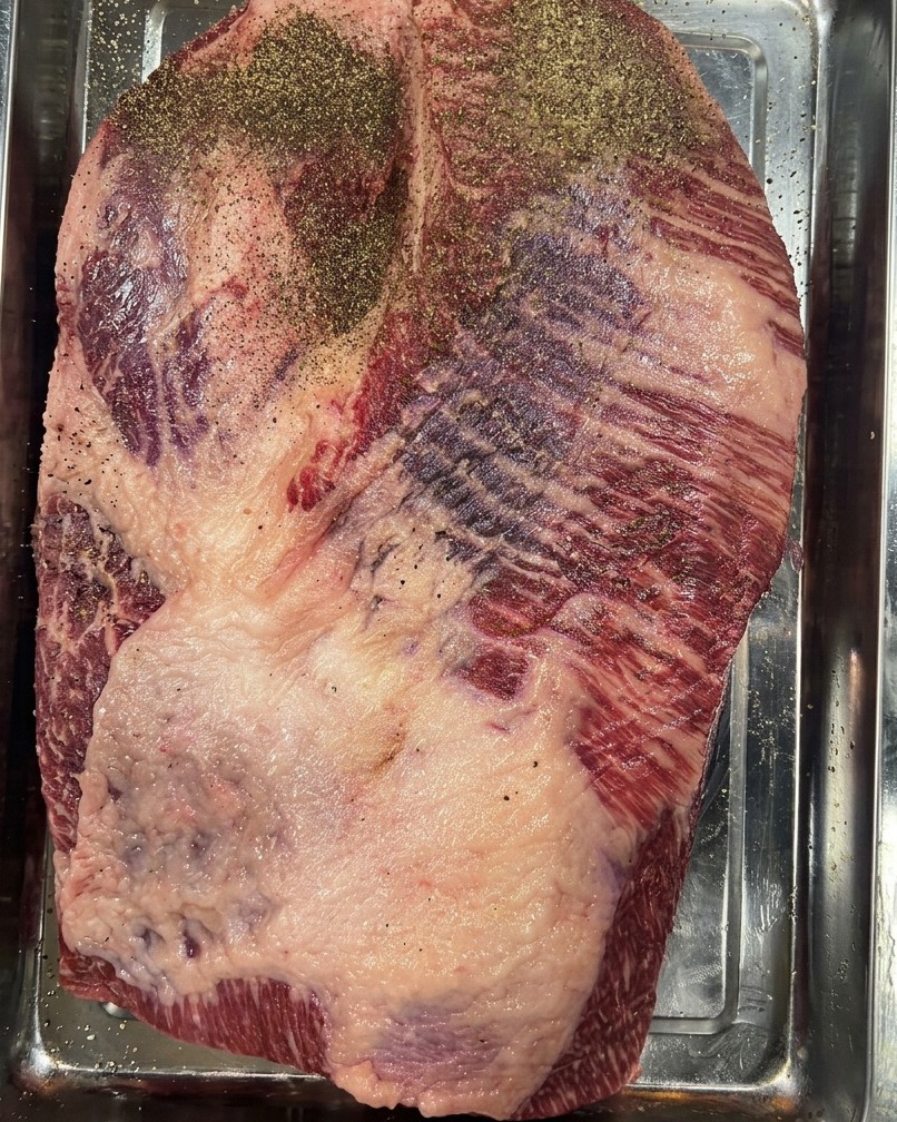 A raw beef brisket resting in a stainless steel tray, heavily seasoned with a layer of ground black pepper.