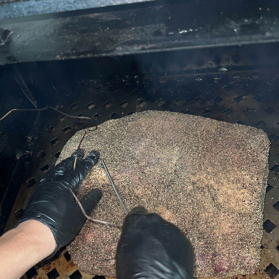 A person wearing black gloves inserting a metal thermometer probe into a seasoned brisket inside a dark smoker.