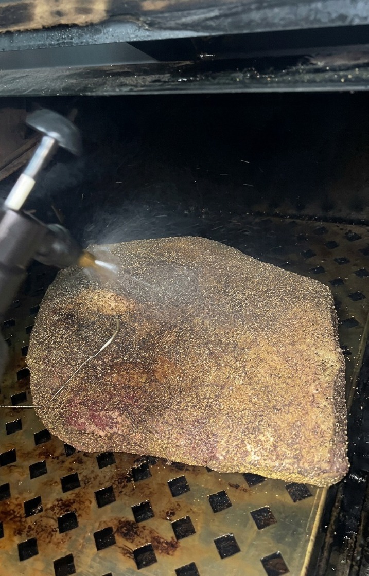 A fine mist of apple cider vinegar being sprayed onto a brisket that is developing a dark bark inside the smoker.