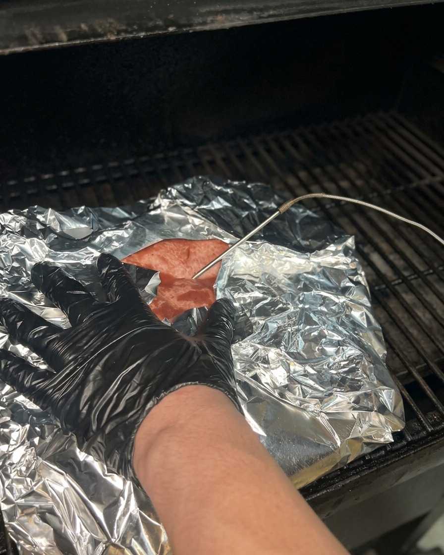 A temperature probe being pushed through aluminum foil into a cooked brisket to check for tenderness and temperature.