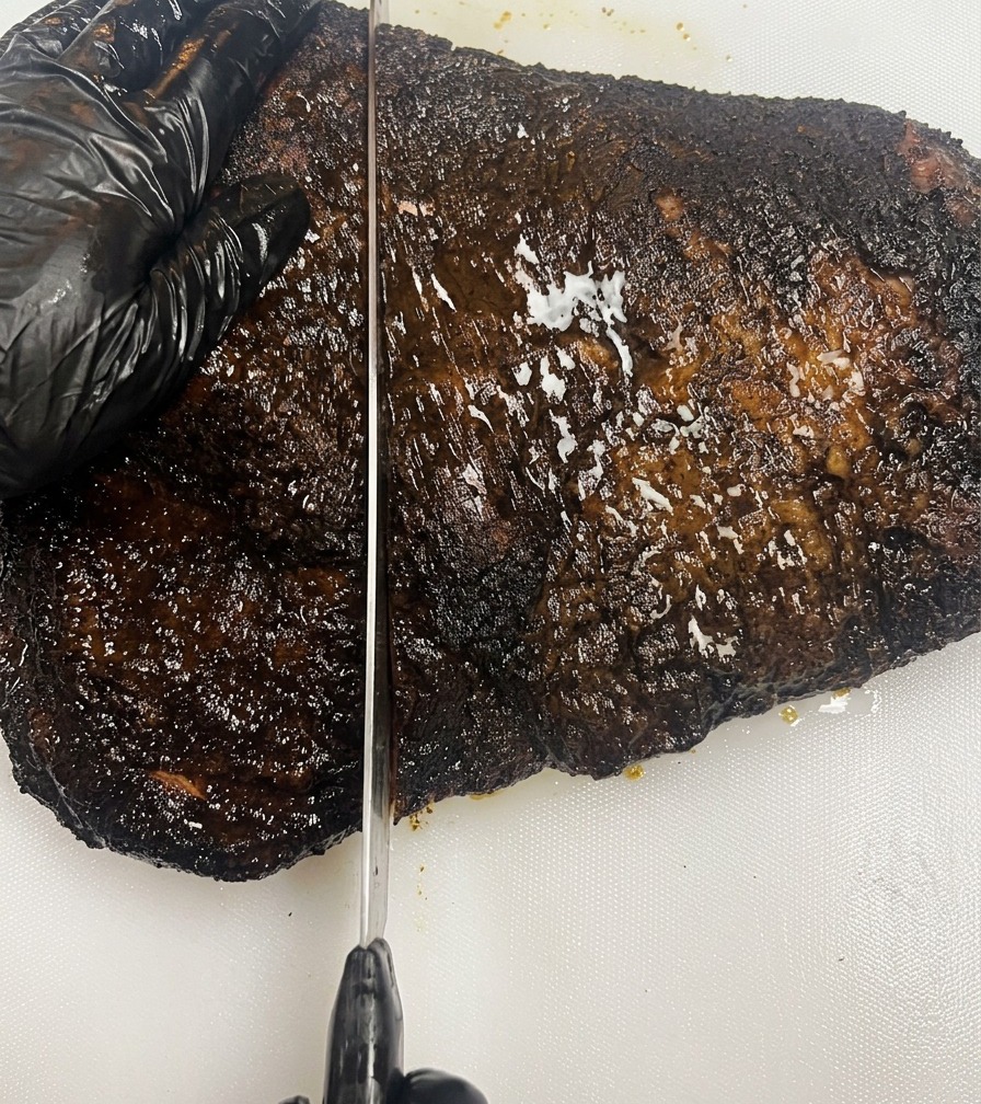 A black-gloved hand steadying a dark, bark-covered smoked brisket on a white cutting board while a long knife slices through the meat.