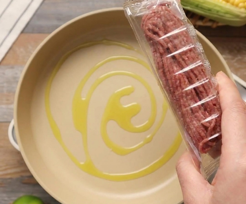 A package of raw ground beef being held over a light-colored pan containing a swirl of olive oil.