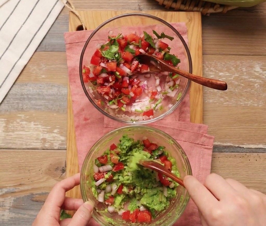 A person using a wooden spoon to mix fresh tomato salsa into a bowl of mashed avocado.