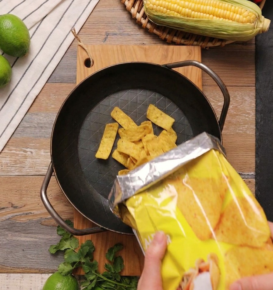 Yellow triangular corn chips being poured from a yellow bag into a heavy black cast-iron skillet on a wooden surface.