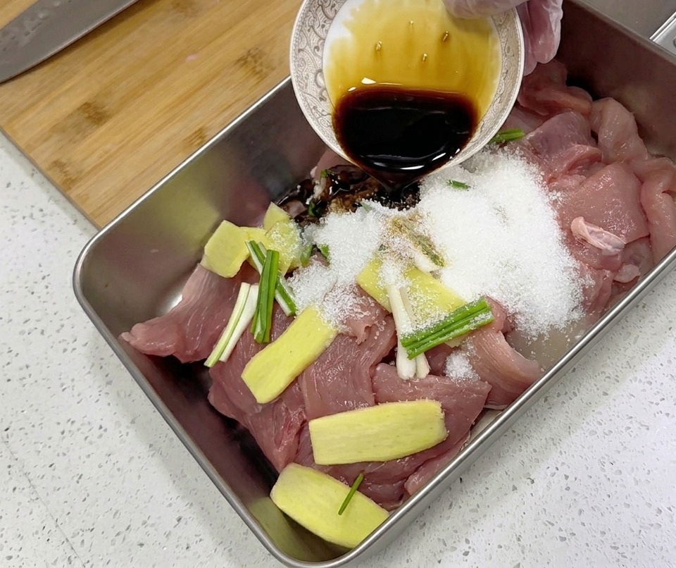 Dark liquid seasoning being poured from a small ornate bowl over raw pork slices, ginger, and green onions in a metal tray.