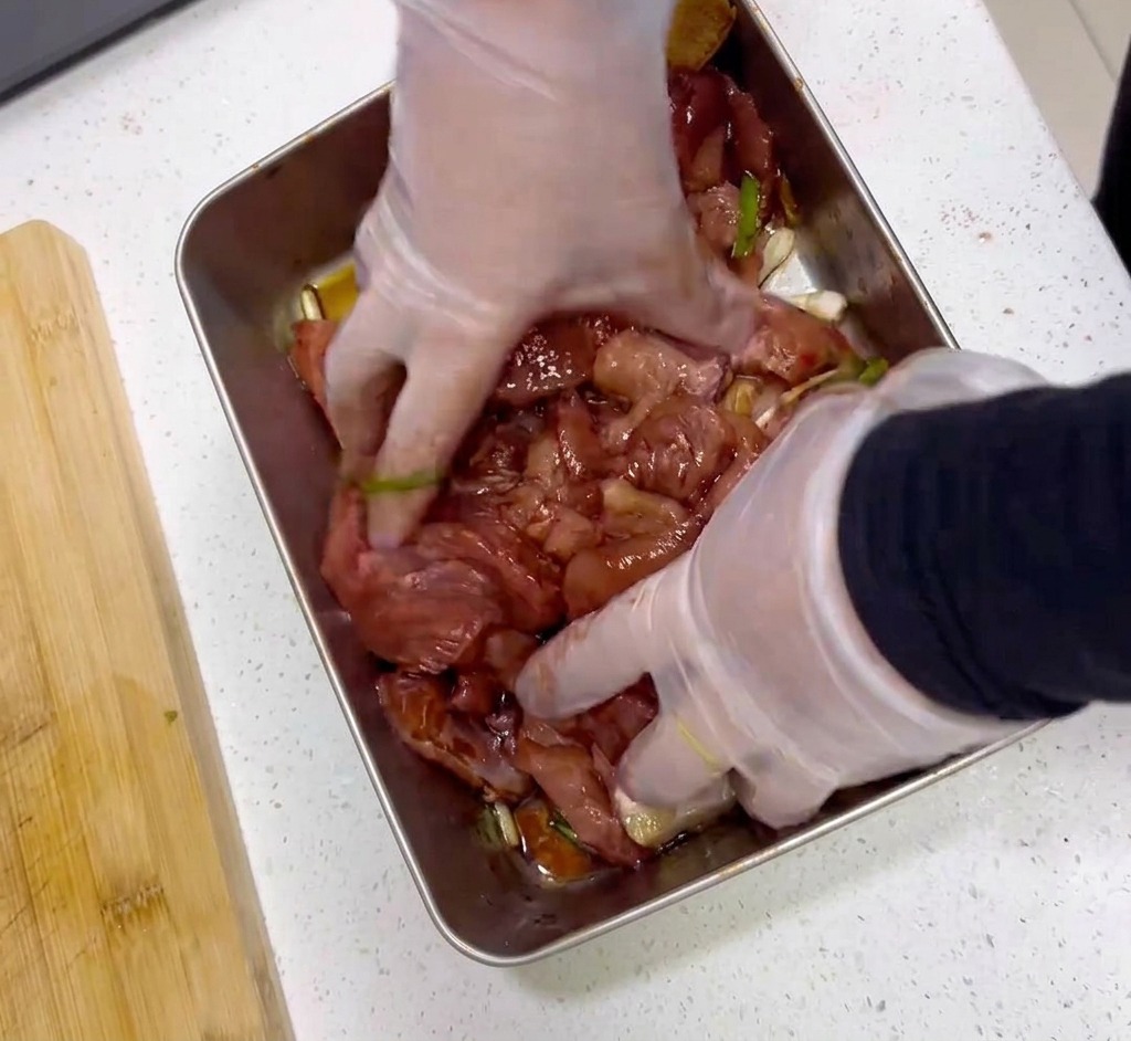 Hands wearing clear plastic gloves mixing slices of raw meat with a dark red marinade in a stainless steel tray.