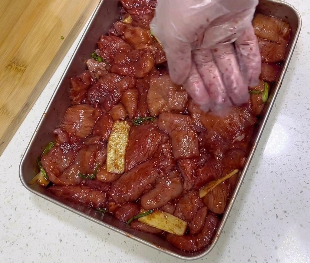 Marinated red pork slices in a stainless steel tray being seasoned with a spoonful of coarse red chili flakes.