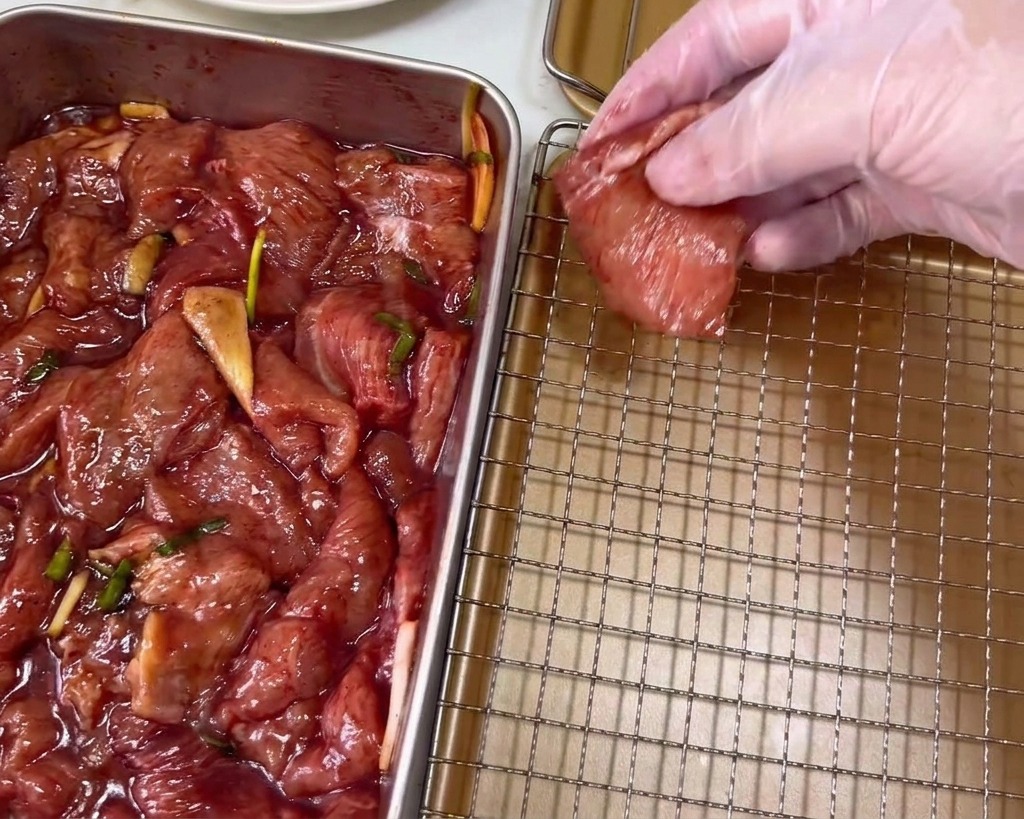 A hand in a clear plastic glove laying a dark red marinated pork slice onto a gold-colored wire rack.