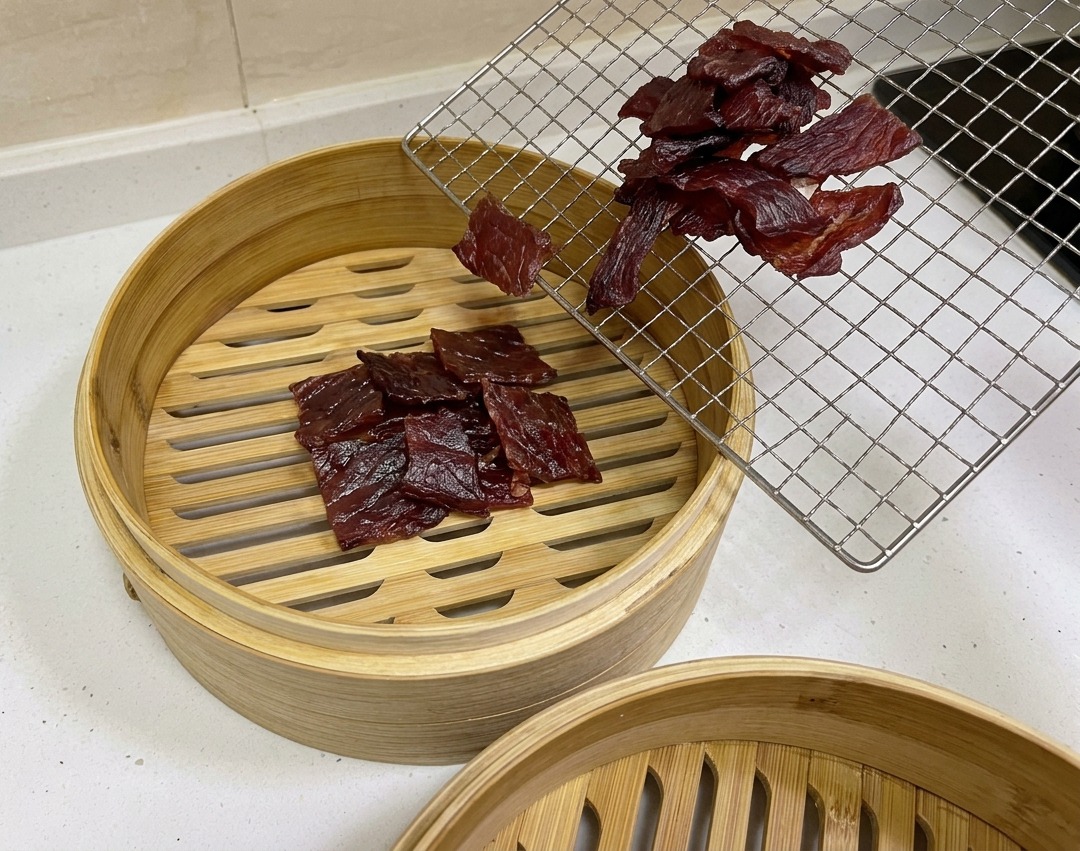 Dark reddish-brown air-dried pork pieces being moved from a wire cooling rack into a circular bamboo steamer basket.