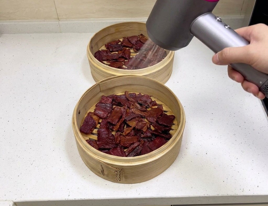 A person holding a modern grey and pink hairdryer, blowing air over pieces of dark brown pork jerky resting inside two bamboo baskets.
