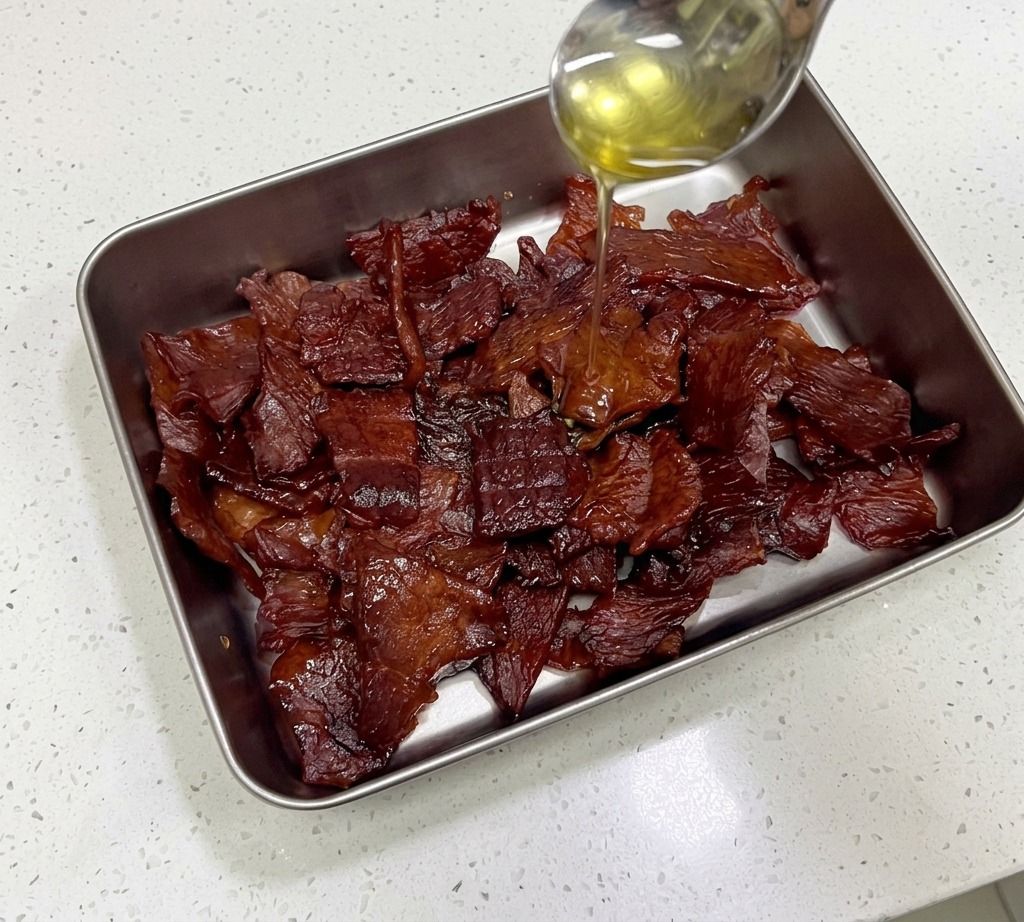 Golden honey being poured from a stainless steel spoon onto a pile of glossy, dark mahogany pork jerky strips in a metal container.