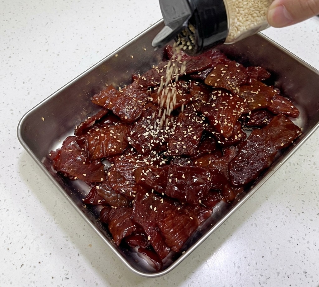 Toasted white sesame seeds falling from a glass shaker onto shiny, dark pork jerky strips in a silver tray.