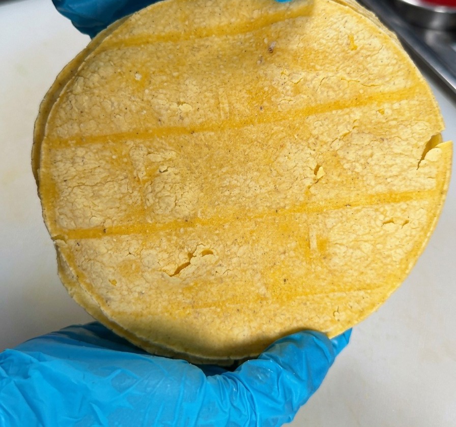 A person wearing blue gloves holding a round stack of yellow corn tortillas over a white kitchen surface.