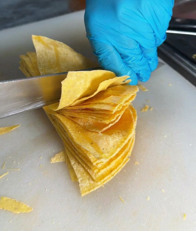 A sharp knife slicing through a stack of corn tortillas to create triangular chip shapes on a white cutting board.