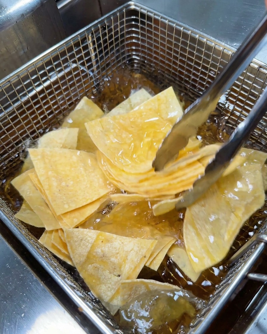 Metal tongs placing a pile of triangular tortilla pieces into a wire mesh deep fryer basket.
