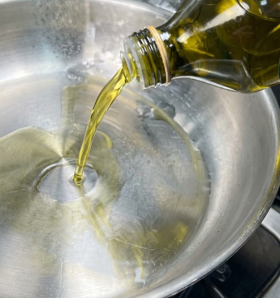 A stream of golden olive oil being poured from a glass bottle into the center of a clean stainless steel frying pan.