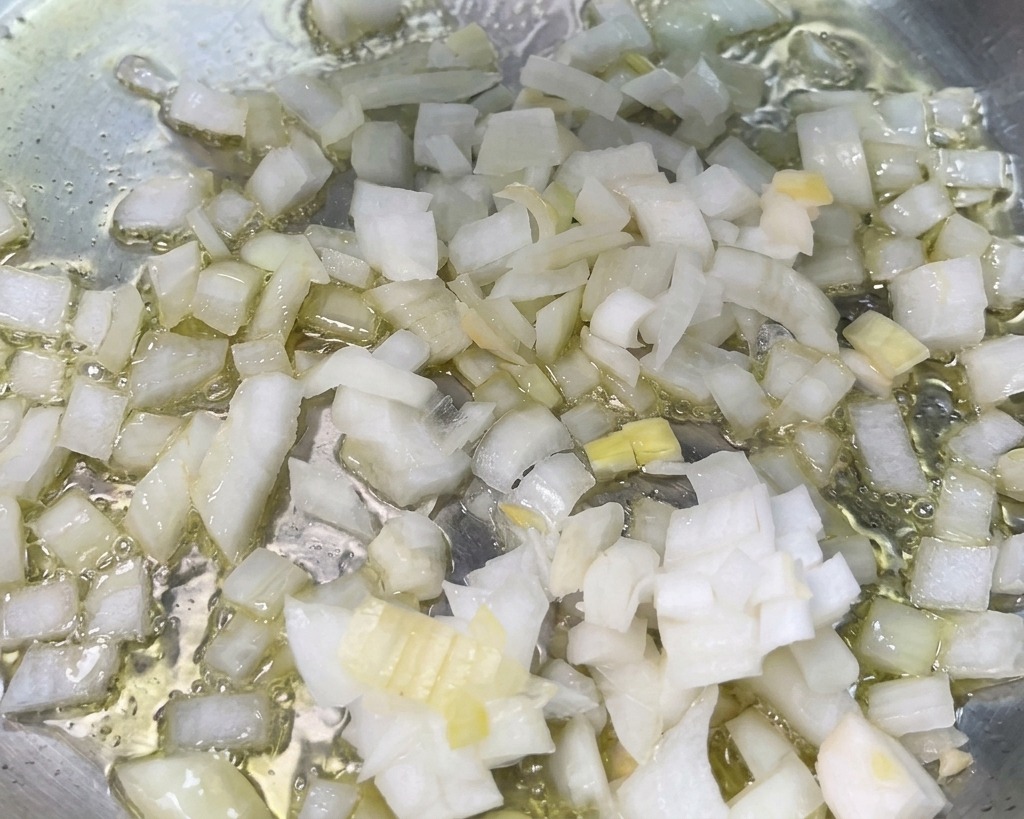 Diced white onions sizzling in hot oil at the bottom of a stainless steel skillet.