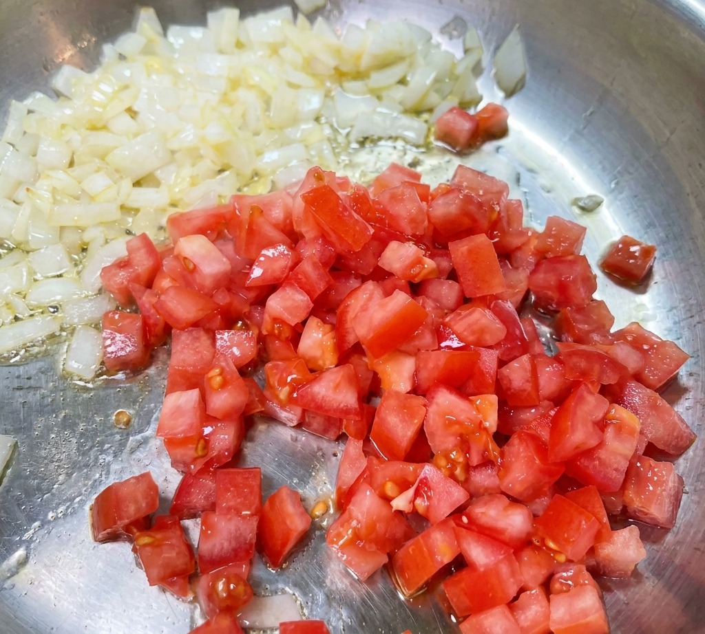 Diced red tomatoes being stirred into a pan of translucent, sautéed onions.