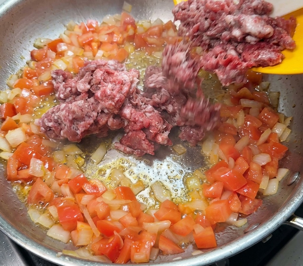 Raw ground beef being added from a yellow plate into a pan containing sautéed onions and tomatoes.