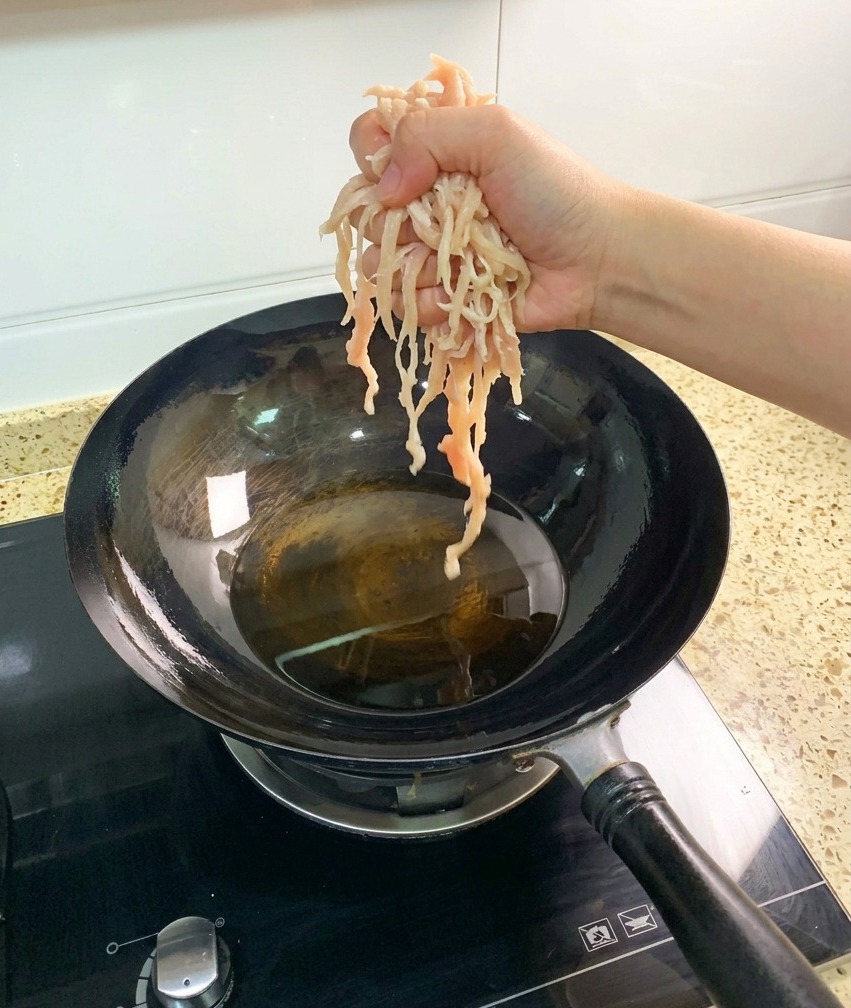 A hand dropping pale, raw shredded chicken breast pieces into a dark wok filled with hot, clear cooking oil.