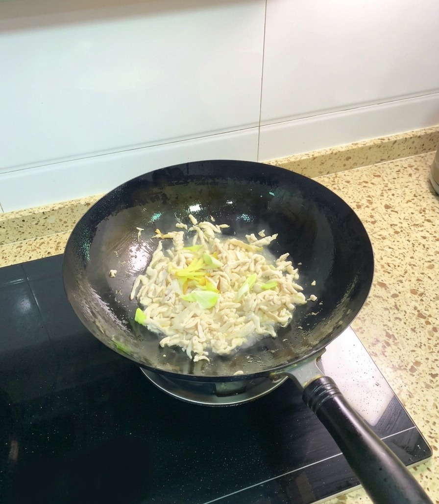 Opaque, partially cooked chicken shreds in a wok being tossed with fresh green onion segments and ginger slices.