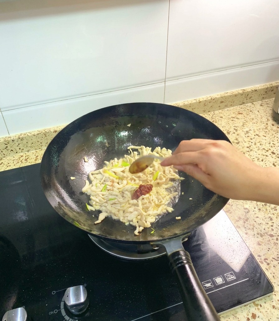 A stainless steel spoon adding a dollop of thick red Pixian broad bean paste to a wok of sautéed chicken and aromatics.