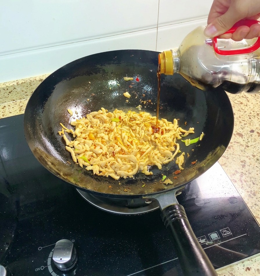 A hand pouring dark soy sauce from a large bottle into a wok containing shredded chicken and green aromatics.