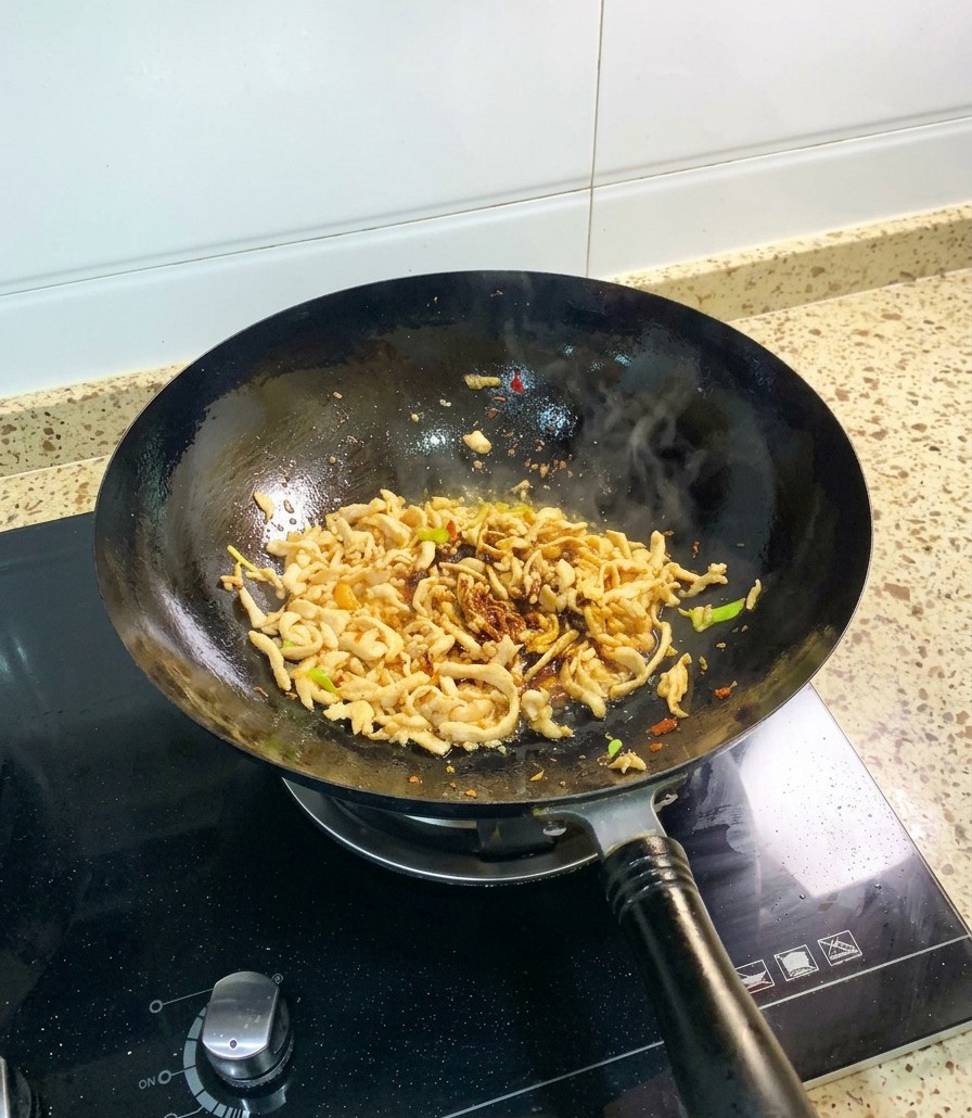 Shredded chicken in a black wok being stir-fried with dark soy sauce and a visible addition of thick oyster sauce.