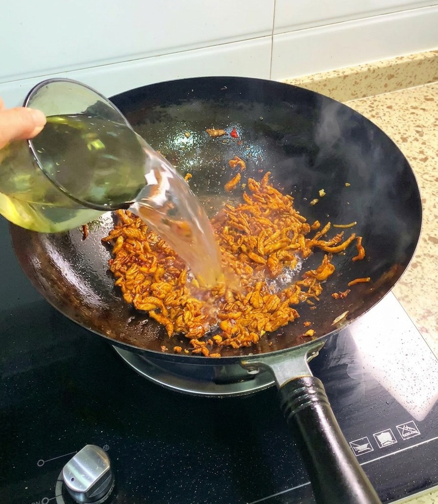 A stream of clear water being poured from a glass cup into a wok filled with seasoned, stir-fried chicken shreds.
