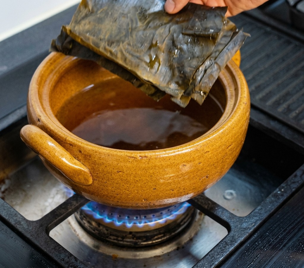A hand placing dark green, rectangular dried kombu sheets into a brown clay pot sitting on a gas stove burner.