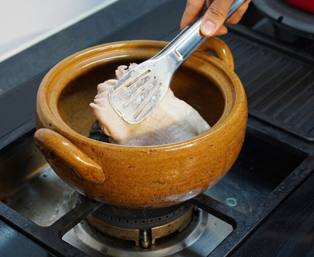 Metal tongs placing a blanched, pale chicken thigh into the brown ceramic pot where the kombu sheets are already visible.