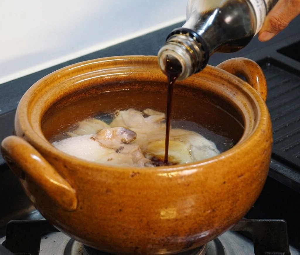 A hand pouring dark liquid seasoning from a glass bottle into the pot containing chicken and water.