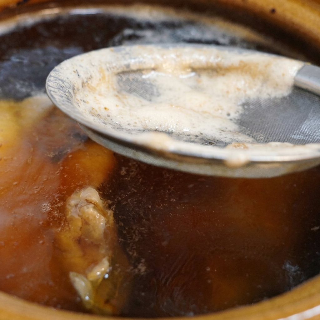 A fine-mesh metal strainer lifting white foam from the surface of a dark, bubbling chicken broth in a ceramic pot.