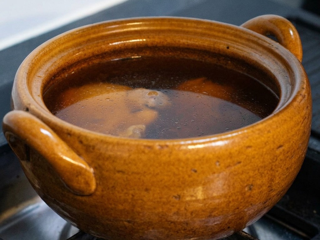 A brown ceramic pot on a gas stove filled with a dark soy-based broth simmering gently, with the lid being partially lifted to reveal the chicken and kombu inside.