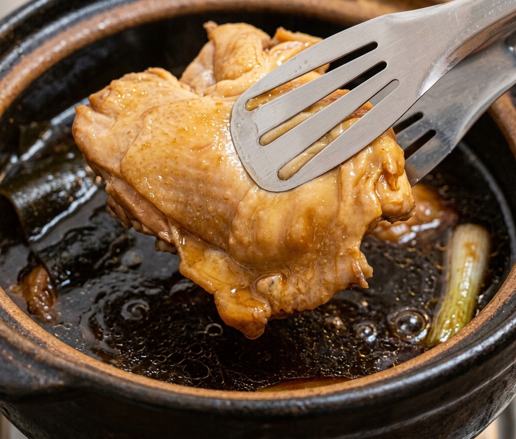 Metal tongs lifting a piece of cooked, browned chicken thigh out of a pot of dark chicken broth.