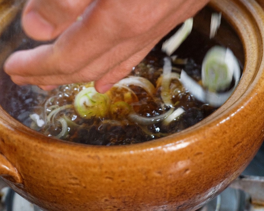 Hands dropping a handful of sliced green scallions into a pot of bubbling dark chicken broth.