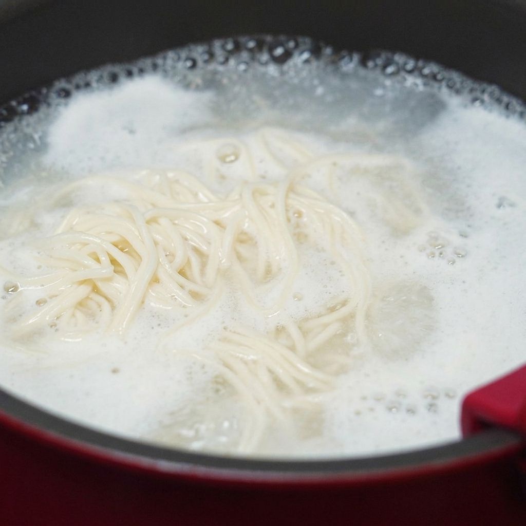 White wheat noodles swirling in a pot of vigorously boiling water with thick starchy foam rising to the edges.