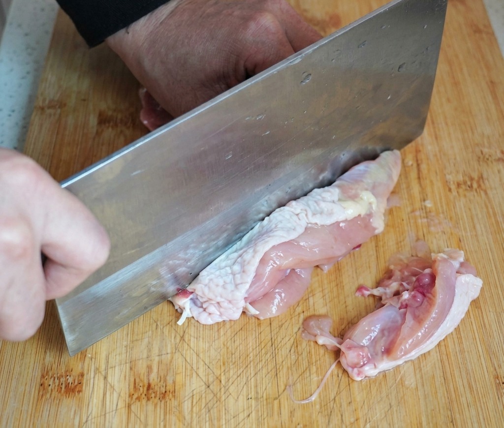 A person using a large metal cleaver to remove the bone from a raw chicken thigh on a wooden cutting board.