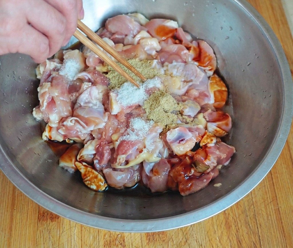 A hand using chopsticks to sprinkle white seasoning over raw chicken chunks in a stainless steel bowl containing dark sauces.