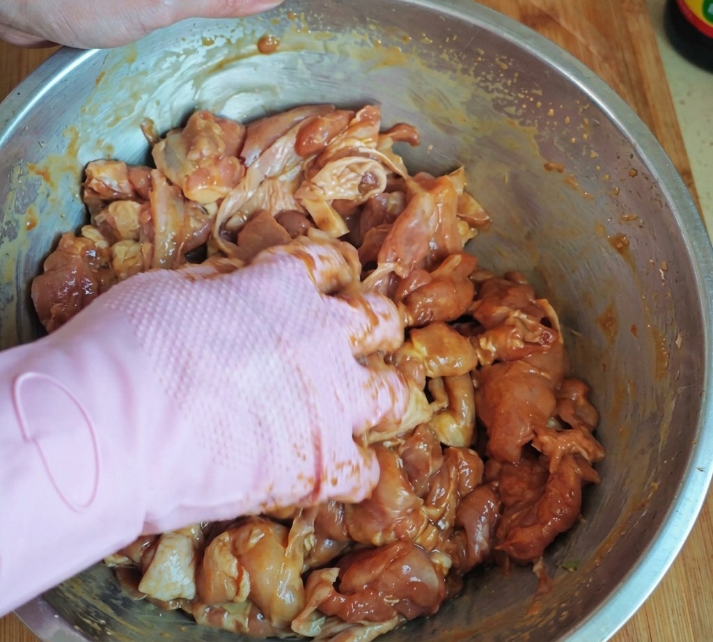 A hand in a pink glove thoroughly mixing marinated chicken pieces with starch and oil in a large stainless steel bowl.
