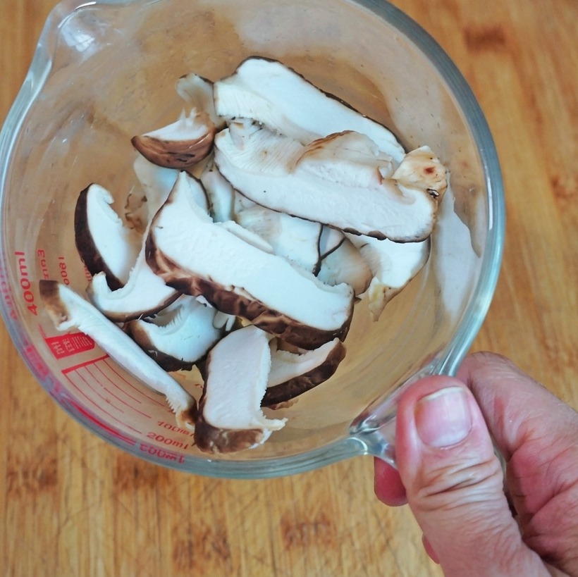 A top-down view of sliced fresh shiitake mushrooms inside a clear glass measuring cup sitting on a wooden board.