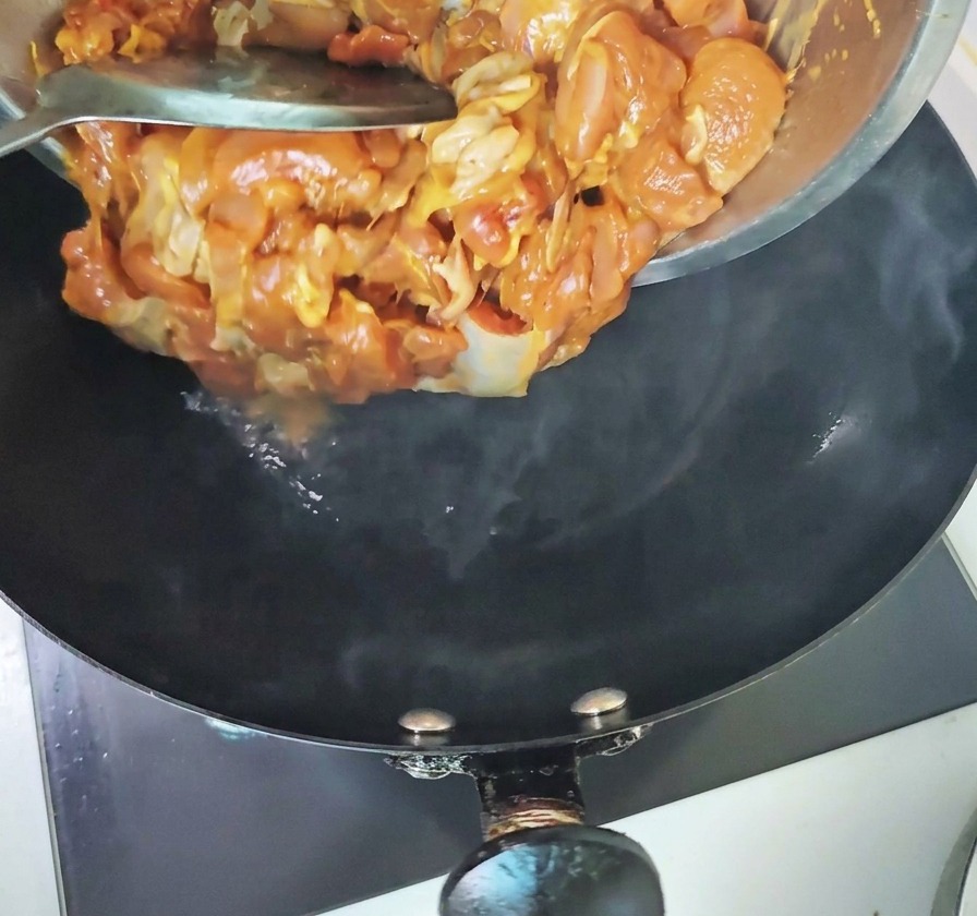 Marinated chicken pieces being poured from a metal bowl into a hot, steaming black wok.