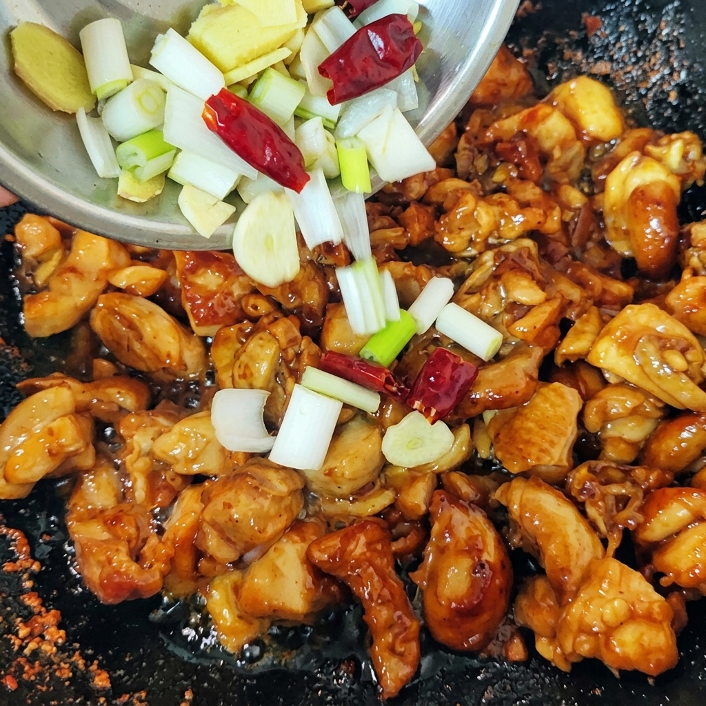 A mix of chopped green scallions, ginger slices, and red dried chilies being poured from a small bowl into a wok filled with seared chicken pieces.