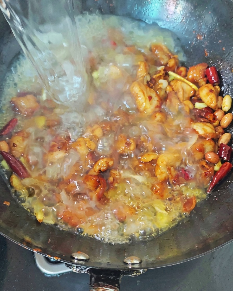 A stream of boiling water being poured into a dark wok containing seared chicken and red chilies, creating a bubbling broth.